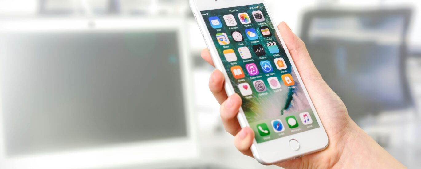 Close-up view of a smartphone displaying apps, held by a hand, with a blurred laptop in the background.